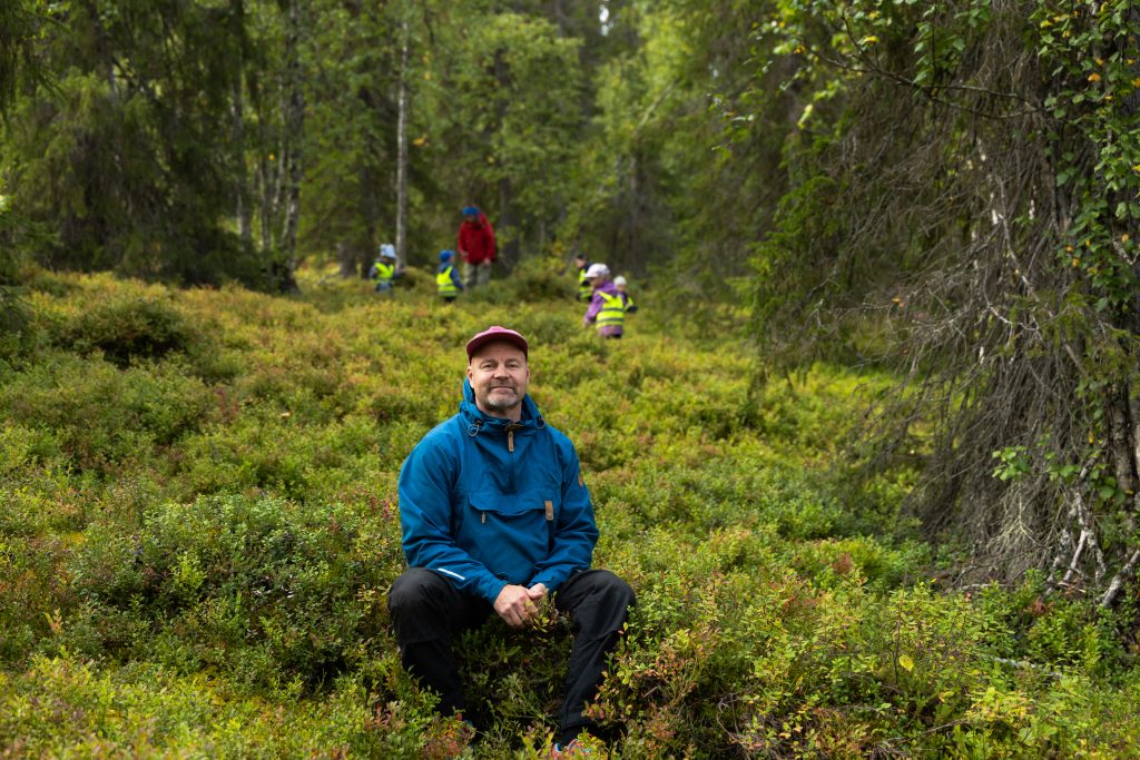 Janne Kylmämaa sits among heather, with a group of children in hi-vis vests on a forest excursion behind him, Äkäslompolo