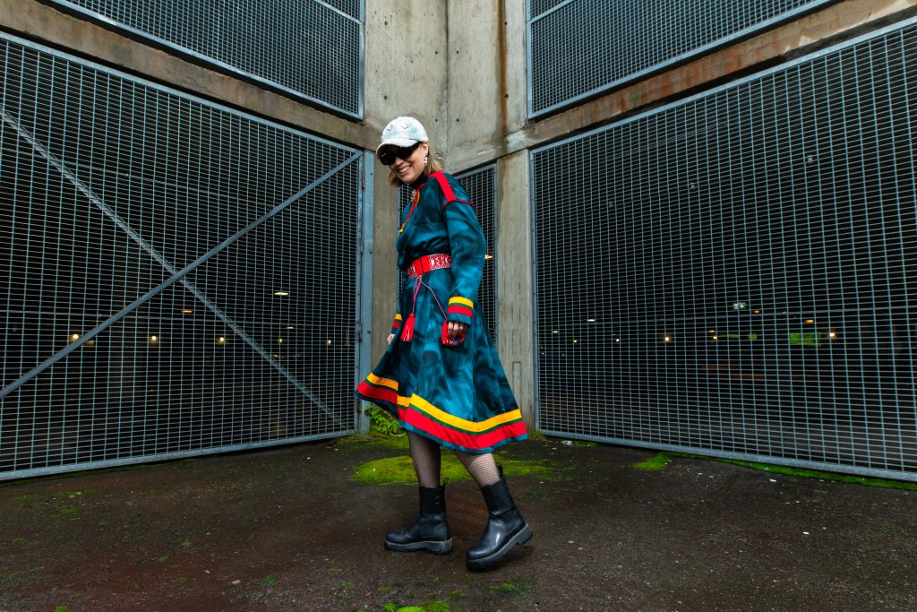 Eleonora Alariesto in a traditional, colourful costume and cap, walking outdoors in front of a wire fence, Rovaniemi