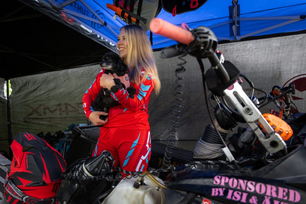 Pinja Paaso holds a small black dog while wearing a red racing suit at a snowmobile event, Ivalo