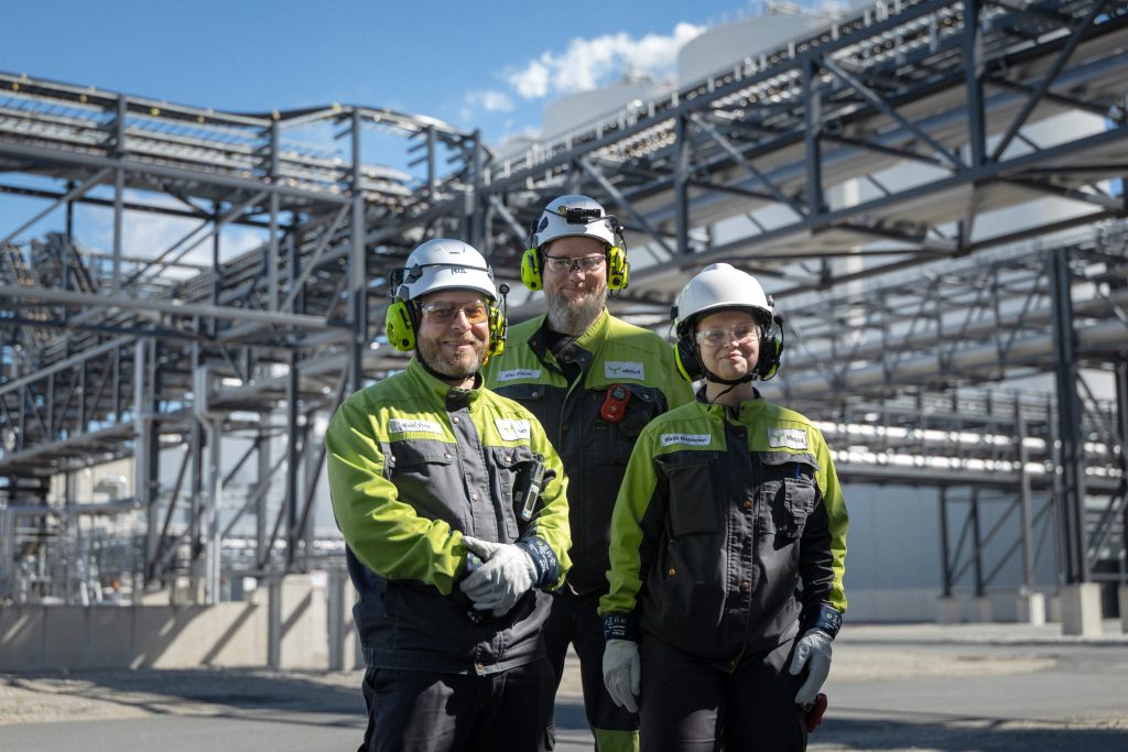 Three workers in hard hats and hi-vis vests pose in the yard of an industrial plant, Kemi