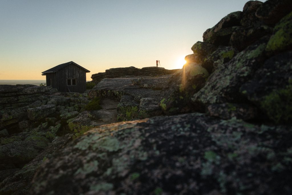 Fell landscape at sunset, an old timber building on the rocks and a photographer silhouetted on the cliff