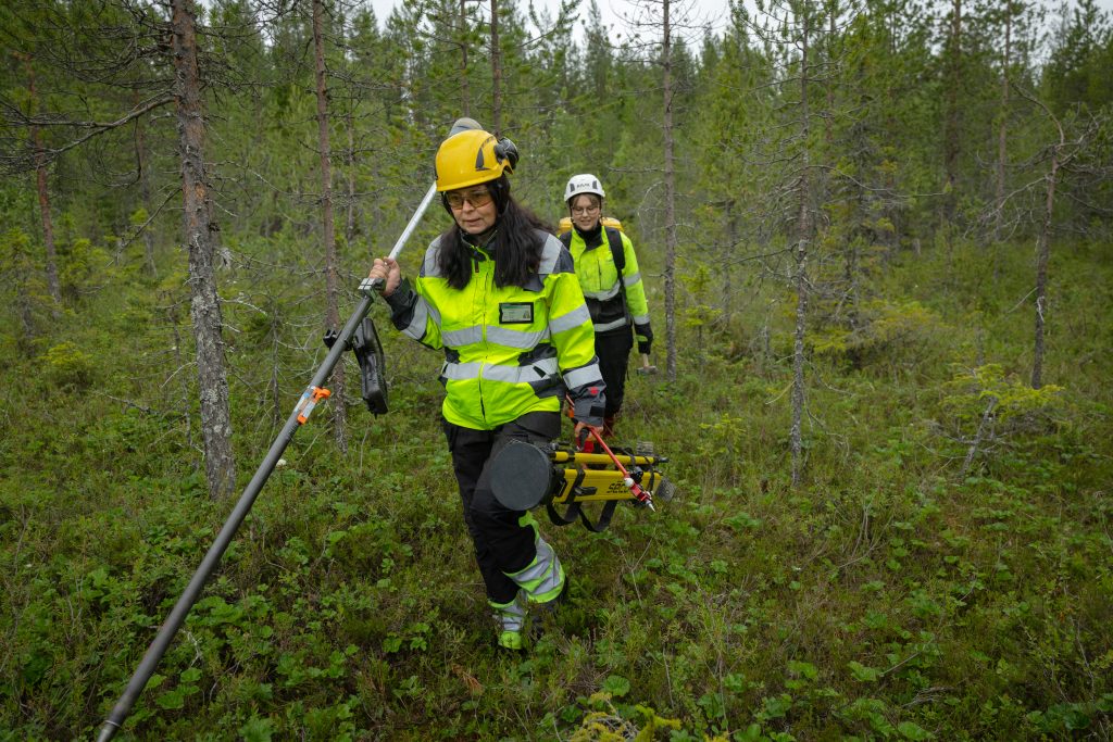 Merja Mustonen and Mira Moberg walk through the forest in protective clothing and helmets, carrying measuring equipment, Rovaniemi
